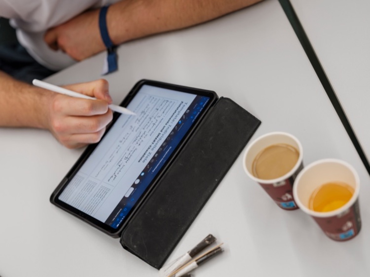 Nahaufnahme einer Person, die sich auf einem Tablet Notizen macht, daneben zwei Kaffeebecher